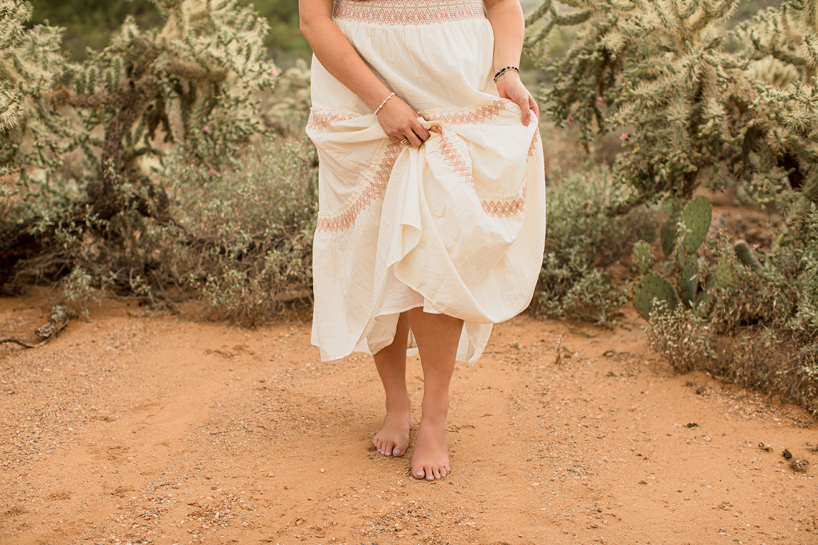 Heather Flynn in a flowing cream dress holding her skirt as she stands barefoot on desert sand among cacti